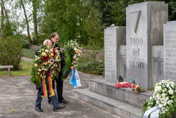 Bild vergr&ouml;&szlig;ern: Zwei M�nner tragen Kr�nze mit Blumen und B�ndern zu einem Gedenkstein auf einem Ehrenfriedhof