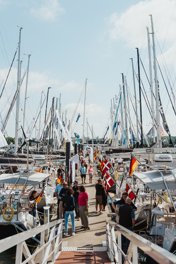 Bild vergr&ouml;&szlig;ern: Menschen schlendern zwischen dicht an dicht liegenden Segelbooten mit Flaggen verschiedener L&auml;nder auf einem Steg beim ancora Yachtfestival 2024