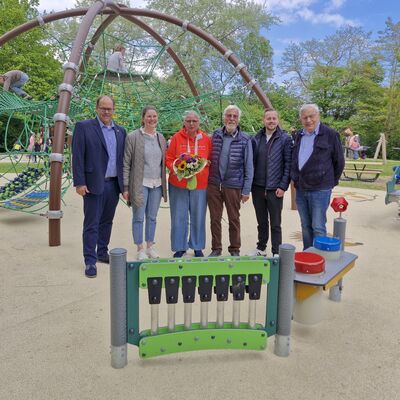 Bild vergr&ouml;&szlig;ern: Sechs Personen stehen vor einem inklusiven Spielplatz mit Kletterger�st und Spielmusikinstrumenten, eine Person h�lt einen Blumenstrau�