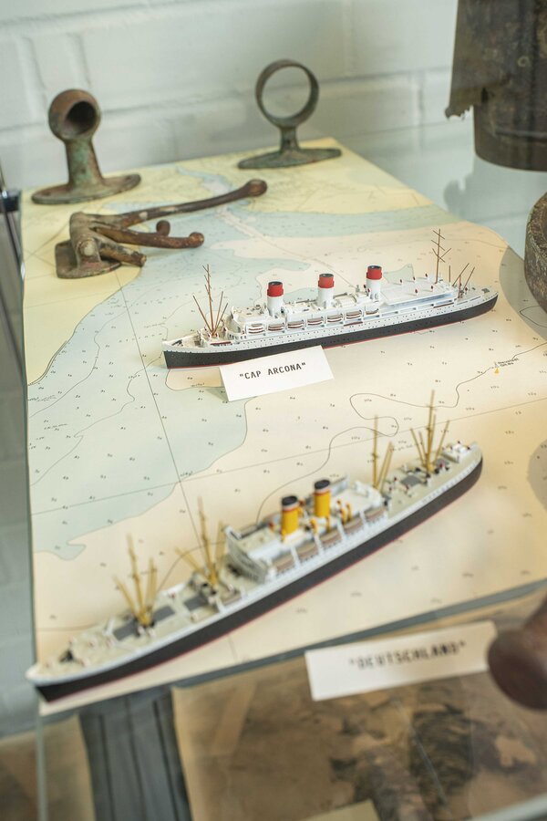 Bild vergr&ouml;&szlig;ern: Zwei Modellschiffe mit den Beschriftungen "Cap Arcona" und "Schleswig-Holstein" auf einer Seekarte mit historischen Ankern im Hintergrund