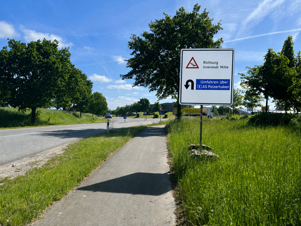 Bild vergr&ouml;&szlig;ern: Verkehrsschild an einer Landstra�e mit Hinweis auf Umleitung �ber AS Pelzerhaken und Richtung Innenstadt Mitte, umgeben von Gras und B�umen unter blauem Himmel