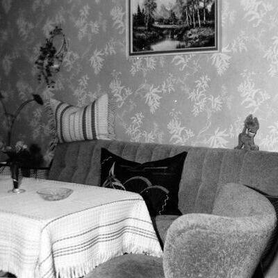 Bild vergr&ouml;&szlig;ern: Wohnzimmer mit gemusterten Tapeten, Sofa mit Kissen, Tisch mit Tischdecke und Vase, Sessel, Stehlampe und Fernseher aus dem Jahr 1961