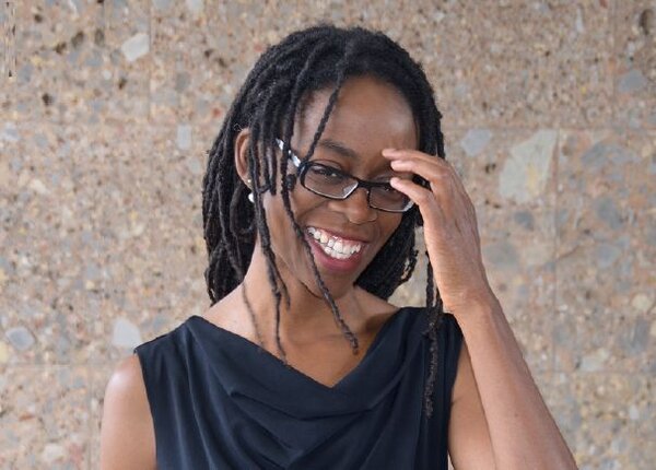 Bild vergr&ouml;&szlig;ern: Sharon Dodua Otoo mit schwarzen Dreadlocks, schwarzer Brille und schwarzem &auml;rmellosem Oberteil l&auml;chelt und ber&uuml;hrt mit der rechten Hand ihre Brille vor einer Steinwand.