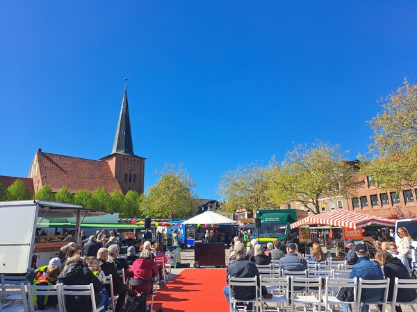 Bild vergr&ouml;&szlig;ern: Menschen sitzen auf Klappst�hlen vor einem Klavier auf einem Marktplatz mit Marktst�nden und einer Kirche im Hintergrund